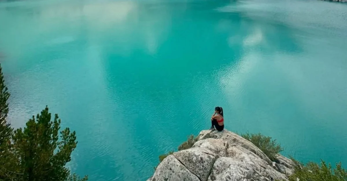 Woman on cliff, turquoise lake, solo journey reflection.