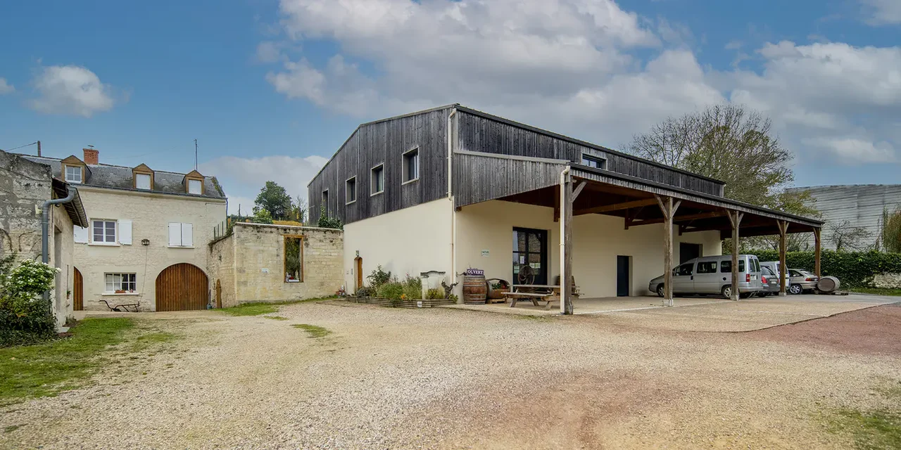 The exterior of a winery building with light-colored walls and a wooden barn-like structure with a covered entrance, parked vehicles nearby.
