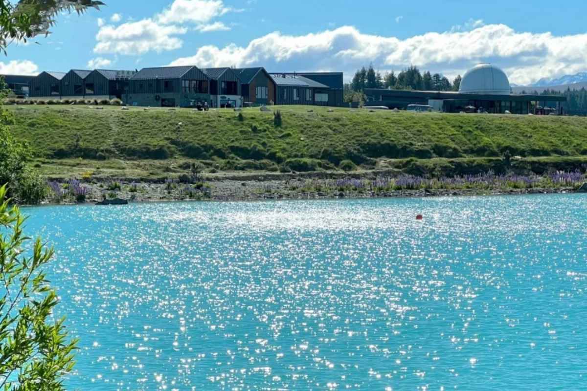 Modern eco-lodge buildings overlooking pristine turquoise lake with rolling green hills and mountains in background