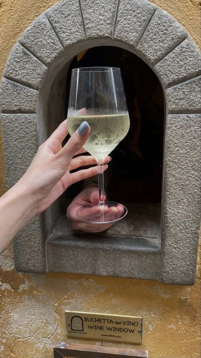 Hands receiving white wine through official Florence wine window with Buchetta del Vino identification plaque