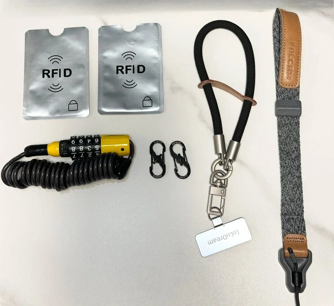 Essential travel security gear including RFID blocking sleeves, cable lock, S-biner clips, wrist lanyard and camera strap on white background