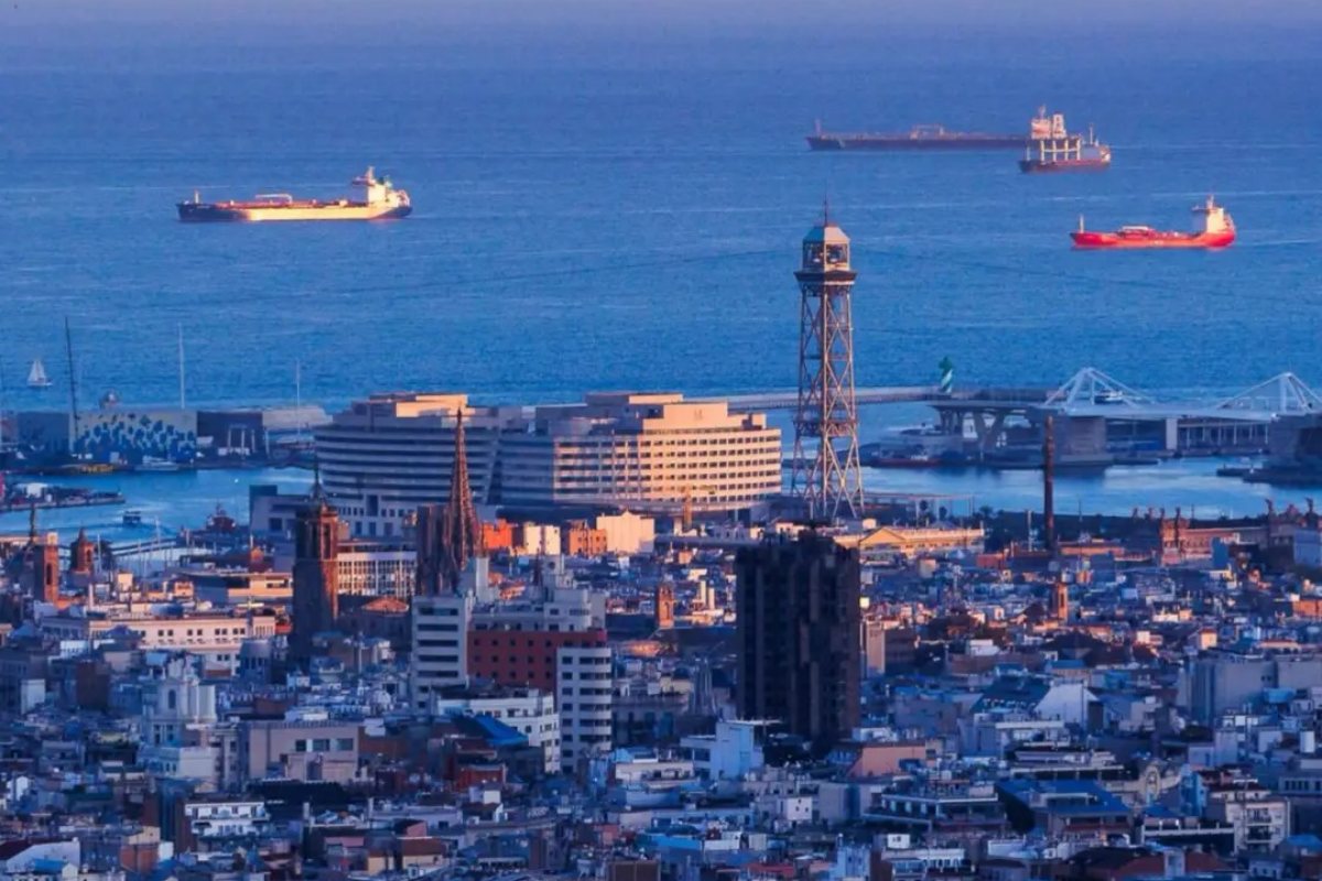 Close-up aerial view of Gaudí's Sagrada Familia cathedral towers glowing in golden sunset light with Barcelona cityscape and Mediterranean Sea backdrop
