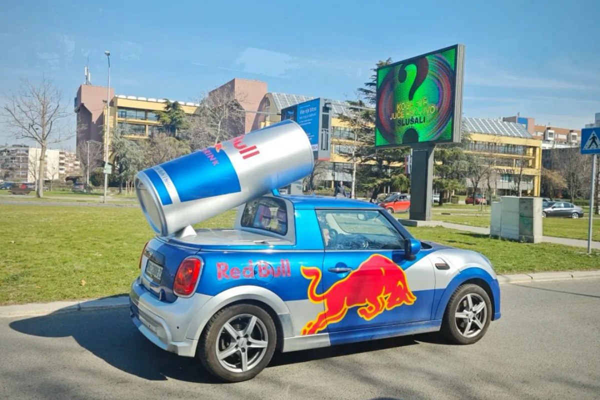 Red Bull branded Mini Cooper with giant energy drink can on roof driving through Niš city center with modern buildings in background